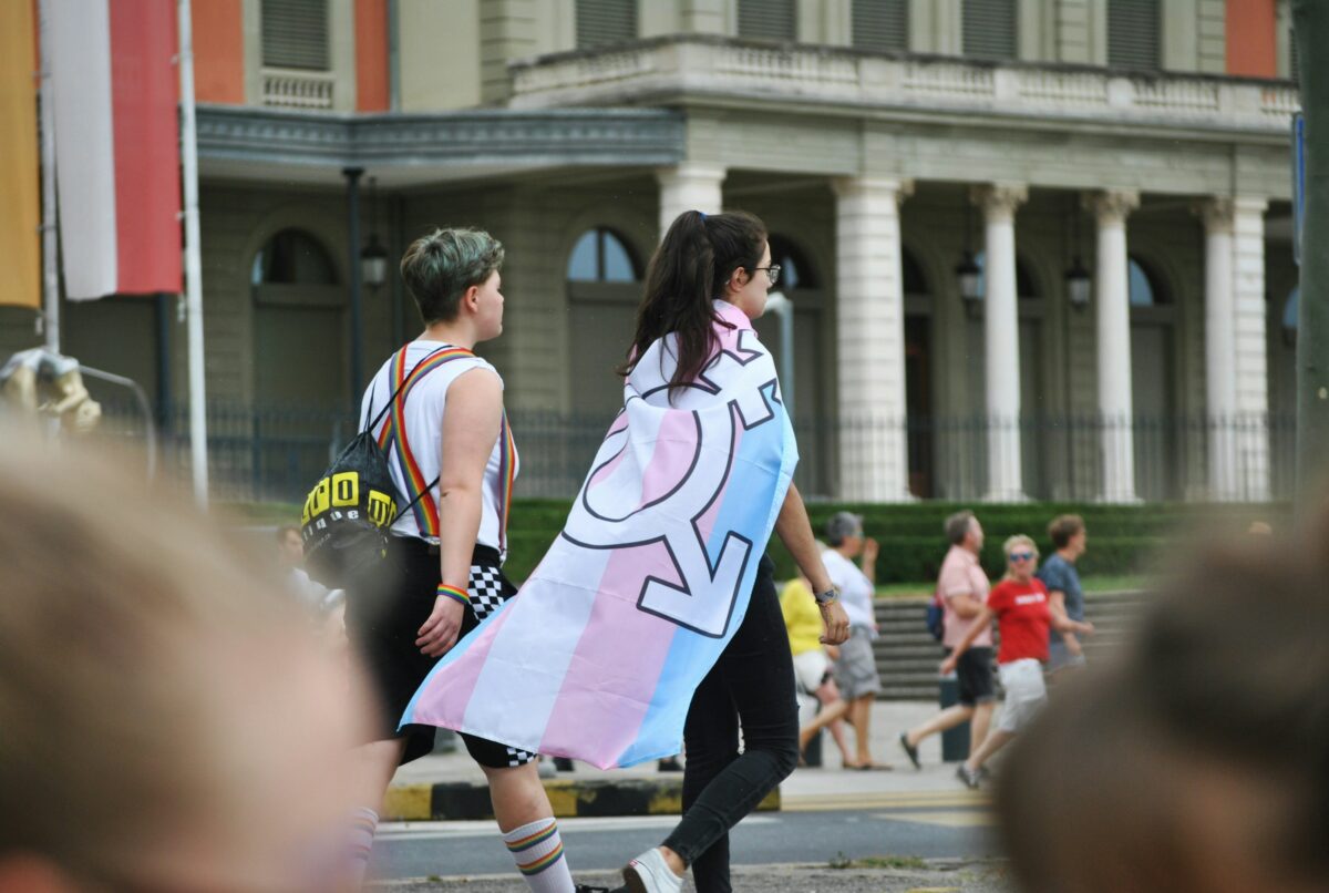 Image prise dans la rue de deux personnes de dos marchant, l'une d'elle avec un drapeau aux couleurs des transidentités comme une cap.